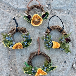 May include: Four small, handcrafted grapevine wreaths. Each wreath features dried orange slices, greenery, and berries. The wreaths have brown leather or black cord loops for hanging. The wreaths are arranged on a textured, gray surface.