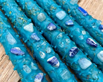 May include: Five teal blue candles with embedded crystals. The candles are decorated with small, clear and blue crystals. The candles are arranged in a row on a wooden surface.