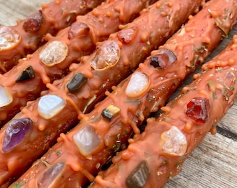 May include: Close-up of five orange handmade candles. Each candle is decorated with various gemstones and dried herbs. The candles are arranged diagonally, showing their unique, textured surfaces and decorative elements.