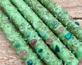 May include: Five green candles with embedded crystals and gold flecks. The candles are arranged in a fan shape, with the tips of the candles pointing towards the right side of the image.