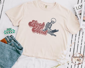 May include: Cream-colored t-shirt featuring a skeleton reading a book, with the words "Book Nerd" in a retro font. The shirt is paired with blue jeans and a green and white checkered hair clip.