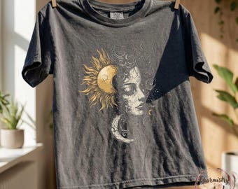 May include: Dark gray t-shirt featuring a celestial design. The graphic includes a sun, moon, and a woman's face with intricate details. The shirt is hanging on a clothesline, showcasing its design and color.