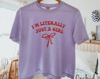 May include: A light purple t-shirt with the text "I'M LITERALLY JUST A GIRL" in red, along with a red bow graphic. The shirt is hanging on a wooden hanger.