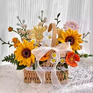 May include: A woven basket filled with handmade crochet flowers. The arrangement includes sunflowers, daisies, and other blooms in shades of yellow, orange, and white. A sheer white ribbon is tied around the basket's handle. The basket is sitting on a white lace tablecloth.