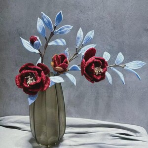May include: A decorative floral arrangement featuring deep red flowers with white centers and light blue leaves. The flowers are arranged in a textured, light brown vase, set against a gray backdrop. The arrangement sits on a white cloth.