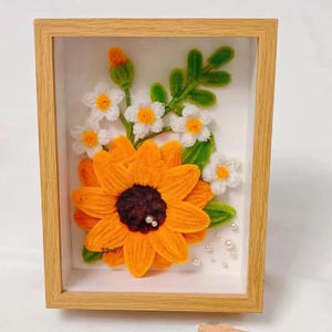 May include: A framed floral art piece featuring a large orange sunflower with a dark brown center and white pearl accents. White and yellow daisies and green leaves are also included. The artwork is encased in a light brown wooden frame.