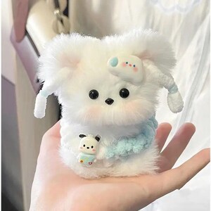 May include: A fluffy white plush toy shaped like a dog with black eyes and a small black nose. The toy has light blue accents, including a bow and a scarf. A smaller toy is held in the dog's paws.