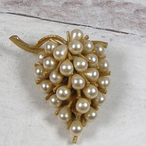 May include: A gold-toned brooch in the shape of a bunch of grapes. The grapes are represented by numerous off-white, pearlescent beads. The brooch has a gold-colored stem and leaf design, creating a classic and elegant accessory.