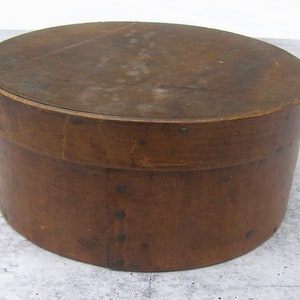 May include: A round, wooden box with a lid. The box is a warm brown color, with visible wood grain and metal rivets. The lid is slightly darker than the body of the box. The box is likely used for storage.
