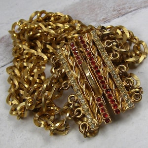 May include: A gold-toned necklace with a decorative clasp. The clasp features rows of red and clear rhinestones set in a gold metal frame. The chain is made of interlocking links.