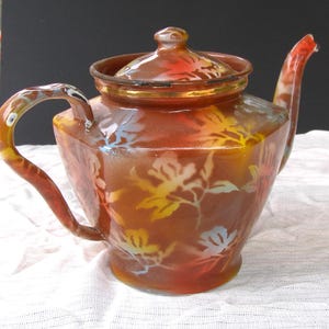May include: A vintage brown teapot with a lid and curved handle. The teapot features a floral pattern in yellow, white, and red. The spout is curved and the handle is arched. The teapot is sitting on a white surface.