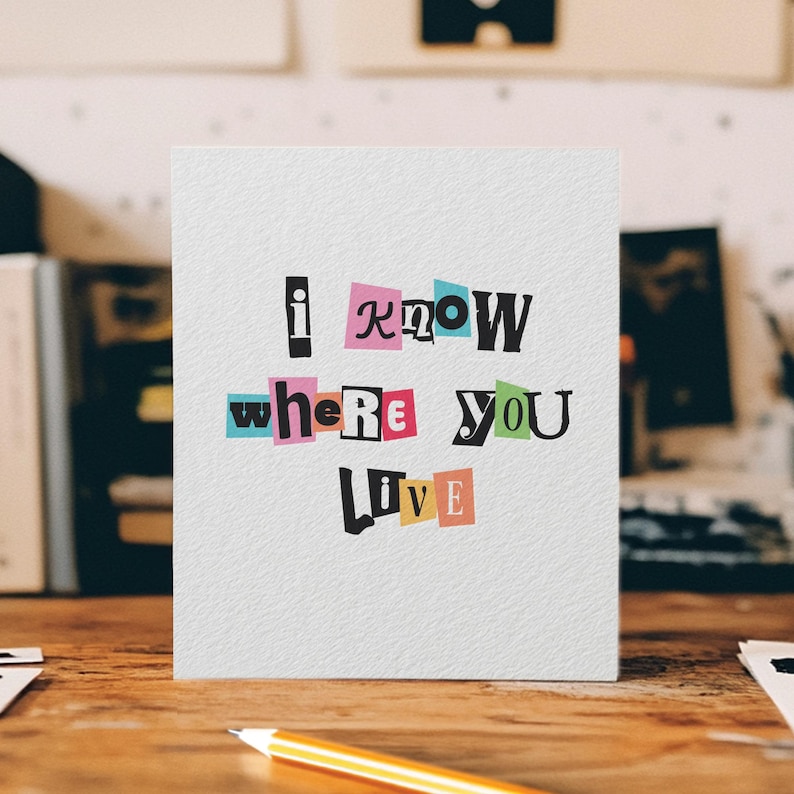 May include: A white greeting card with the text "I know where you live" in a cut-out letter style. The words are in various colors, including pink, green, and yellow. The card is on a wooden surface with a pencil.