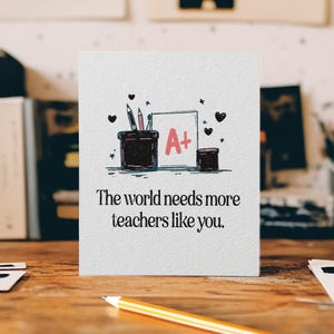 May include: A white greeting card with the text "The world needs more teachers like you." The card features a drawing of pencils, a grade of A+, and hearts. A yellow pencil rests on the wooden surface.