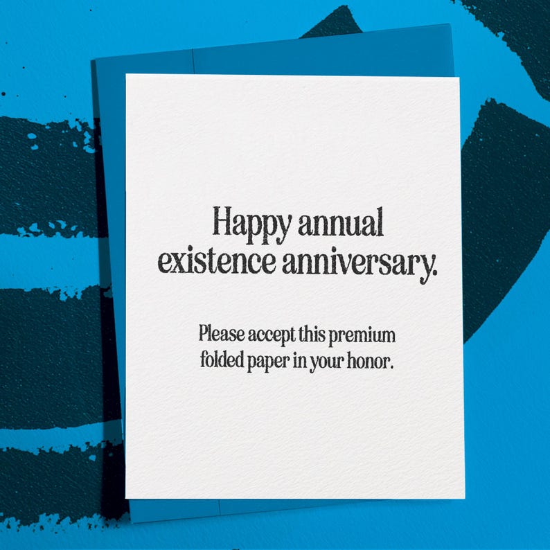 Puede incluir: Una tarjeta de felicitaci&oacute;n blanca con el texto "Happy annual existence anniversary." y "Please accept this premium folded paper in your honor." La tarjeta est&aacute; dentro de un sobre azul, sobre un fondo azul.