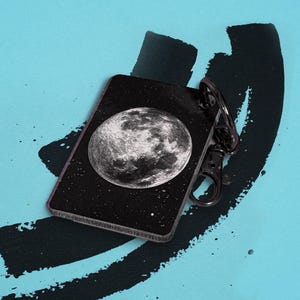 May include: A black rectangular keychain featuring a detailed grayscale image of the moon against a starry background. The keychain has a silver-colored clasp and is set against a teal background with black brushstrokes.