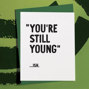 May include: A white greeting card with the text "YOU'RE STILL YOUNG" in bold black letters, followed by "...ISH." The card is inside a green envelope, set against a green background with dark green brushstrokes. A humorous birthday card.