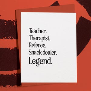 May include: A white greeting card with the words "Teacher. Therapist. Referee. Snack dealer. Legend." in black font. The card is set against a red background with darker red abstract shapes.