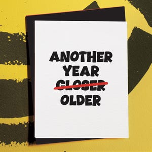 May include: A white greeting card with the text "ANOTHER YEAR CLOSER OLDER" in bold black letters. The word "CLOSER" is crossed out with a red line. The card is on a black envelope with a red border, set against a yellow background.
