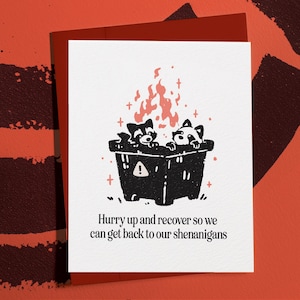 May include: A greeting card with a humorous illustration of two raccoons in a burning trash can. The card has the text "Hurry up and recover so we can get back to our shenanigans" printed in black font on a white background. The card is on a red background.