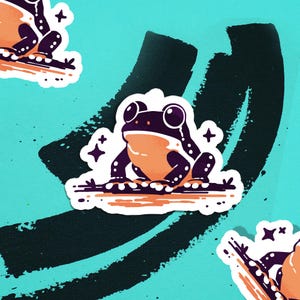 May include: A sticker featuring a cartoon frog with a purple body, orange belly, and large white eyes. The frog is sitting on a brown line, with a teal background and black brush strokes. Decorative star shapes are around the frog.