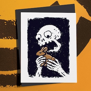 May include: A black and white art print featuring a skull with wide eyes and a gaping mouth, seemingly exhaling a brown rabbit. The rabbit is held by skeletal hands. The print is on a white card, set against a black background.
