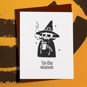 May include: Greeting card with a black and white illustration of a skeleton wearing a witch hat and cloak, holding a cup with a straw. The text "'tis the season" is below the illustration. The card is on a brown background.