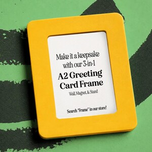 Puede incluir: Un marco amarillo para tarjetas de felicitaci&oacute;n A2 con interior blanco. El texto en el marco dice "Make it a keepsake with our 3-in-1 A2 Greeting Card Frame Wall, Magnet, & Stand." El marco est&aacute; sobre un fondo verde con pinceladas negras.