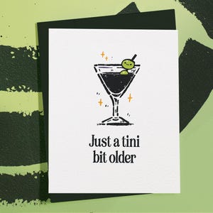 May include: A black and white illustration of a martini glass with two green olives and the text "Just a tini bit older".
