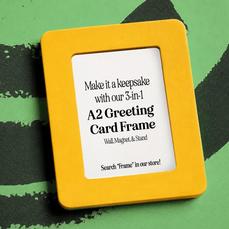 May include: A yellow A2 greeting card frame with a white interior. Text on the frame reads "Make it a keepsake with our 3-in-1 A2 Greeting Card Frame Wall, Magnet, & Stand." The frame is on a green background with black brushstrokes.