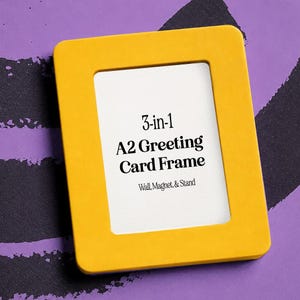 May include: A rectangular yellow picture frame with rounded corners. The white insert features black text that reads 3-in-1 A2 Greeting Card Frame, Wall, Magnet, & Stand. The frame has a smooth, solid-colored border surrounding the central white area.