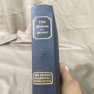 May include: A dark blue hardcover book spine with gold lettering. The top label reads THE NABOB DAUDET, and the bottom label reads THE FRENCH CLASSICAL ROMANCES. A small gold fleur-de-lis emblem is embossed in the center of the textured fabric cover.