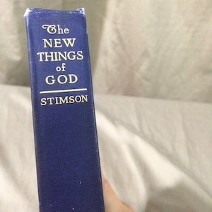 May include: A dark blue book spine with gold lettering that reads The NEW THINGS of GOD and STIMSON. The top edge of the spine shows slight wear and fraying on the corners of the blue fabric cover, while the rest of the spine remains intact.