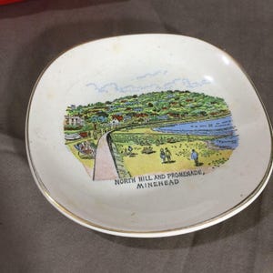 May include: A white ceramic dish with a gold-toned rim featuring a colorful illustration of a coastal landscape. The scene depicts a beach, a promenade, and a hillside with buildings. The text reads NORTH HILL AND PROMENADE, MINEHEAD.