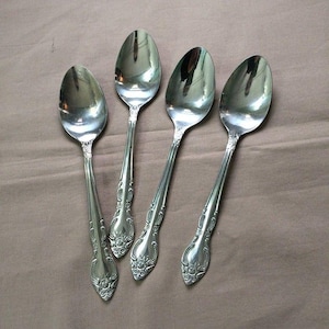 May include: Four silver spoons with ornate floral detailing on the handles. The spoons have a reflective, polished surface, and are arranged on a neutral-toned background. The spoons are a classic style.