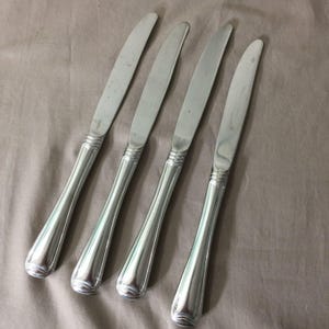 May include: Four silver-colored knives with rounded handles and a decorative band near the blade. The blades are slightly curved and appear to be made of stainless steel. The knives are arranged diagonally on a neutral-toned surface.