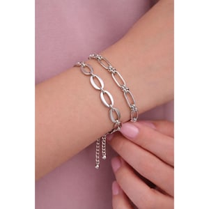 May include: Two silver bracelets are shown on a wrist. One bracelet features oval links, while the other has a chain-like design. The bracelets are made of a shiny, metallic material. The background is a solid pink color.
