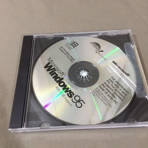 May include: A Microsoft Windows 95 CD-ROM in a clear plastic case. The CD has the Windows logo and text, "With USB Support." The case is black with a ribbed edge. The CD also has the text "CD Sampler" and "Free Games."