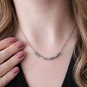 May include: A silver necklace with a series of small, round beads and cylindrical bars. The necklace is worn around the neck and hangs just below the collarbone. The background is a person's skin and clothing.
