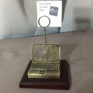 May include: A bronze-colored laptop-shaped photo holder on a dark brown wooden base. The holder has a wire loop to hold a photo or memo. The text "Place photo or memo here" is visible.