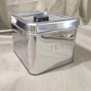 May include: A square, polished stainless steel tea canister with a reflective surface. The container features a removable lid with a small, round, black knob handle centered on top. The word TEA is embossed in capital letters on the front side.