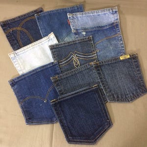 May include: A collection of denim jean pockets in various shades of blue. Some pockets feature decorative stitching in gold or white thread. The pockets are arranged on a neutral-colored surface, showcasing the different washes and styles.