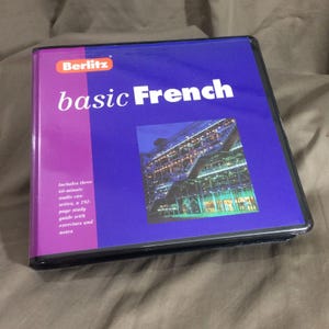 May include: A Berlitz Basic French language learning binder with a purple and blue cover. The cover features the text "Berlitz" and "basic French" alongside a photograph of a modern building. It includes three 60-minute audio cassettes and a 192-page study guide with exercises and notes.
