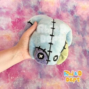 May include: A light blue plush toy with black stitching and patches of purple and green. The toy has a stitched-on button eye and a cross-stitch design. The toy is held in a hand against a colourful background.