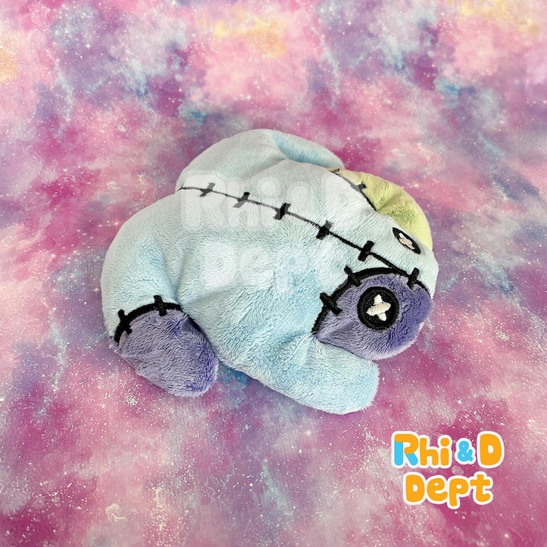 May include: A light blue plush toy with purple details, designed to look like a character with stitched features. Black thread is used for the seams and facial features. The toy has a button eye and the text "Rhi&D Dept" is visible.