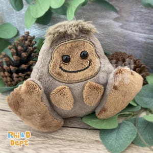 May include: A soft Bigfoot toy with a smiling face, brown fur, and embroidered details. The toy has a light brown body, darker brown arms and feet, and a tuft of brown hair. The toy is on a wooden surface with greenery and pinecones.