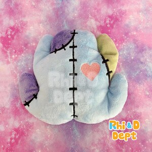 May include: A heart-shaped plush cushion in pastel shades. It features light blue, purple, and yellow sections, with black stitching and a pink heart appliqu&eacute;. The design has a whimsical, handmade appearance, reminiscent of a patchwork creation.