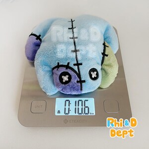 May include: A light blue plush toy in the shape of a stitched-together creature, with purple and green accents. The toy is on a digital scale, which reads 0:10.6 lb:oz. The toy has black stitched details and button eyes.