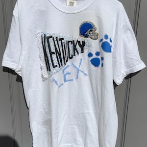 May include: White t-shirt with a blue and black patch that says "Kentucky" and a blue and gold football helmet patch.