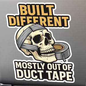 May include: A sticker on a laptop featuring a skull with duct tape around its head and a roll of duct tape. The text reads "BUILT DIFFERENT" above the skull and "MOSTLY OUT OF DUCT TAPE" below.