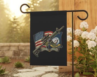 Puede incluir: Bandera de jardín negra con un rifle superpuesto a la bandera estadounidense y un emblema naval. La bandera dice "HOIST THE COLOURS" en texto blanco. La bandera se muestra en un soporte de metal negro en un jardín.